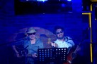 Dwóch mężczyzn grających na gitarach i śpiewających. Mają założone ciemne okulary i kapelusze. Całość oświetlona jest niebieskim światłem. 