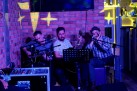 Na zdjęciu trzech mężczyzn. Dwóch z nich gra na gitarach. Widać sprzęt muzyczny oraz część baru. Światła są przyciemnione, jedynie emitowane są przez lampę jasne gwiazdy.
