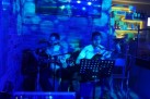 Dwóch mężczyzn grających na gitarach i śpiewających. Widać sprzęt muzyczny, ekran wiszący na ścianie oraz część baru. Całość oświetlona jest niebieskim światłem. 
