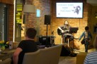 Na zdjęciu widać występ muzyczny w kameralnym wnętrzu. Główne elementy zdjęcia to: kobieta siedząca na krześle, grająca na gitarze akustycznej i śpiewająca do mikrofonu, chłopiec stojący po jej prawej stronie, obok statywu z nutami, fragment publiczności w pierwszym rzędzie, z widocznym mężczyzną siedzącym na kanapie. W tle, na ceglanej ścianie, wisi duży telewizor. Wyświetla on napis 'ROMUALD KUBIK PAPIER I PRZESTRZEŃ' oraz grafikę przedstawiającą głowę barana. Wokół sceny znajdują się głośniki i sprzęt nagłośnieniowy.
