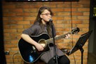 Zdjęcie przedstawia młodą dziewczynę, która gra na gitarze i śpiewa do mikrofonu. Za nią znajduje się ceglasta ściana, zaś przed nią stoi stojak na nuty. 