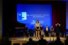 Młoda kobieta ubrana w białą bluzkę i czarną spódniczkę występuje. Za nią inni uczestnicy wydarzenia. Widać także jury. 