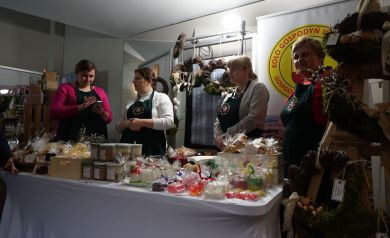 Cztery kobiety z koła gospodyń wiejskich w jednakowych fartuchach. Przed nimi i za nimi wystawiane przedmioty i plakat koła gospodyń wiejskich. 