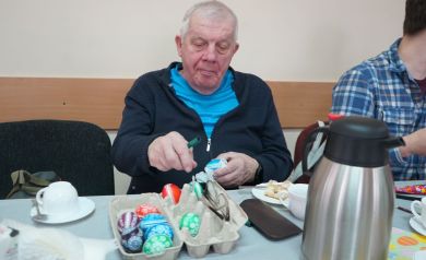 Pan w niebieskiej koszulce na wierzchu z ciemną bluzą ozdabiający kroszonki. Obok niego siedzi mężczyzna w koszuli w kratkę. Na stole stoi termos, talerzyk z kołaczem, filiżanki, materiały do ozdabiania jaj i kroszonki. 