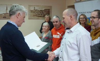Mężczyzna w ciemnym ubraniu wręcza dyplom uczestnikowi. Na drugim planie widać innych uczestników wydarzenia. 