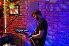 Na zdjęciu muzyk grający na gitarze. Wokół niego inne instrumenty i sprzęt muzyczny. 