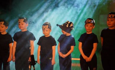 Uczestnicy wydarzenia stojący na scenie, ubrani na czarno z maskami jeży na głowie. Za nimi kłęby ciężkiego dymu.