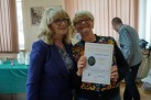 Na zdjęciu dwie kobiety. Obie mają blond włosy i okulary. Jedna z kobiet trzyma w dłoniach dyplom. 