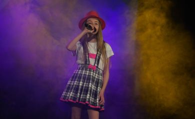 Dziewczynka obrana w spódnicę w kratkę, białą bluzkę i kapelusz stoi na scenie i trzyma mikrofon, do którego śpiewa. Za nią światła rzucające niebieskie i żółte tło oraz kłęby ciężkiego dymu.
