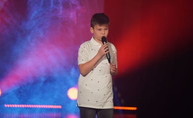 Na scenie chłopiec śpiewający do mikrofonu. Za nim kolorowe światła. 