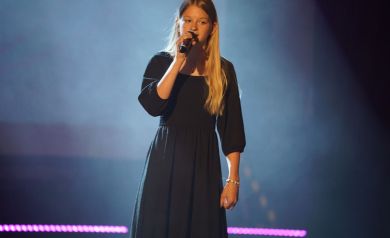 Młoda dziewczyna, o długich blond włosach, ubrana w długą, czarną sukienkę stoi na scenie i śpiewa do mikrofonu. 