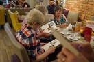 Uczestnicy wydarzenia siedzą przy stolikach. Dwie kobiety przeglądają prawdopodobnie ulotkę związaną z odbywającą się imprezą. 