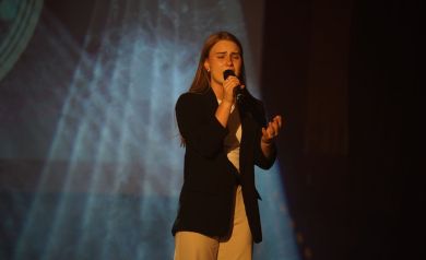 Dziewczyna o długich, blond włosach stojąca na scenie, ubrana w czarny żakiet i jasne spodnie śpiewa do mikrofonu. 