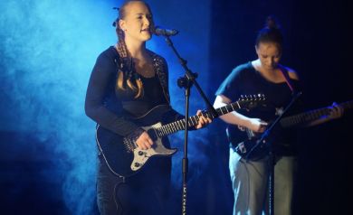 Dwie dziewczyny grające na gitarach, jedna śpiewa do mikrofonu. W tle niebieskie światło i kłęby ciężkiego dymu dodające klimatu. 