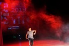 Na zdjęciu znajduje się osoba podczas swojego występu. W tle widać czerwone światło i ciężki dym oraz część wyświetlanego plakatu związanego  wydarzeniem. 