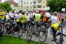 Na zdjęciu grupa rowerzystów zebrana przy rowerach. Mają stroje sportowe, a na głowach założone kaski. Na drugim planie wiać innych uczestników wydarzenia oraz orkiestrę dętą. 