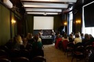 Uczestnicy wyjazdu siedzący na krzesłach w małej sali widowiskowej. O scenę opiera się dwóch mężczyzn.