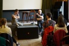Na zdjęciu uczestnicy wyjazdu siedzący na krzesłach oraz dwaj mężczyźni siedzący na scenie. Mężczyzna z wąsem zwraca się do publiki.