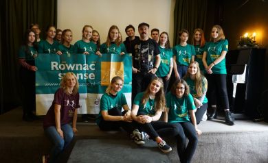 Na zdjęciu znajduje się grupa młodzieży i dorosłych pozująca  wspólnie do zdjęcia na scenie w zamkniętym pomieszczeniu. Większość osób ubrana jest w zielone koszulki z logo programu „Równać Szanse”, a przed nimi rozciągnięty jest baner projektu. W centrum grupy stoi mężczyzna w czarnej koszulce i czapce.  - powiększ