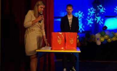 Po prawej kobieta przemawiająca, na środku mężczyzna ubrany na czarno stojący przy stoliku z nagrodami od Strzeleckiego Ośrodka Kultury w Strzelcach Opolskich 