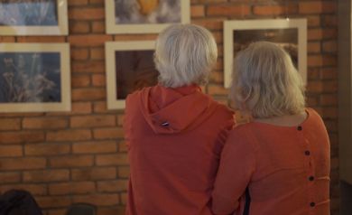 Dwójka uczestników wydarzenia, odwróceni tyłem, ubrani w pomarańczowe bluzy. Spoglądają na obrazy w ramkach powieszone na ceglanej ścianie. 