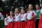 Na zdjęciu dzieci występujące. Stroje dziewczynek to czerwone spódnice, białe bluzki i czerwone korale, w włosach wplątane mają czerwone kokardy, zaś chłopiec widoczny na zdjęciu ma założone ciemne spodnie, białą koszulę oraz zieloną kamizelkę. 
