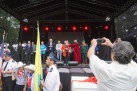 Na zdjęciu widać uroczystość na scenie plenerowej, gdzie grupa osób w różnych strojach – zarówno współczesnych, jak i historycznych – uczestniczy w oficjalnym wydarzeniu. Mężczyzna dokumentuje ten moment aparatem fotograficznym. 
