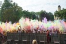 Scena podrzucania przez tłum ludzi w górę kolorowych proszków ,,Holi'. W tle widać ruiny zamku oraz drzewa. 