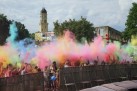 Scena podrzucania przez tłum ludzi w górę kolorowych proszków ,,Holi'. W tle widać ruiny zamku oraz drzewa. 