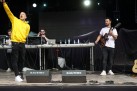 Na zdjęciu czterech mężczyzn. Jeden zwraca się do publiczności. Inny gra na gitarze. Widać również sprzęt muzyczny. 