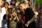 Na zdjęciu widać mężczyznę z tatuażami, który pozuje do zdjęcia z grupą fanów stojących za metalowym ogrodzeniem, podczas gdy dzieci i młodzież entuzjastycznie robią zdjęcia telefonami.