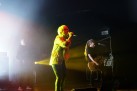 Na zdjęciu kobieta śpiewająca do mikrofonu. W tle dwaj mężczyźni grający na gitarach. Scena jest przyciemniona, a oświetla ją żółte światło. 
