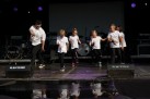 Na scenie grupa taneczna ubrana w białe koszulki i czarne spodnie podczas swojego występu. 
