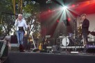 Muzycy na scenie podczas występu. Widać również sprzęt muzyczny i czerwone światło emitowane przez lampy przymocowane na scenie. 