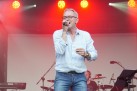 Mężczyzna w błękitnej koszuli śpiewa do mikrofonu. W tle perkusista. Scenę oświetla czerwone światło. 