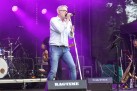 Na scenie mężczyzna śpiewający do mikrofonu. Na drugim planie mężczyzna grający na gitarze oraz sprzęt muzyczny. Całość oświetla fioletowe światło. 