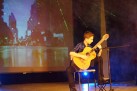 Na zdjęciu chłopak siedzący na krześle i podpierający prawą nogę o podnóżek, ubrany w koszulę w granatowo-czarną kratę gra na gitarze. Przed nim mikrofon. 