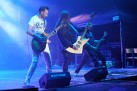 Na scenie trzej gitarzyści podczas swojego występu. W tle widać namiot z sprzętem muzycznym. Przed muzykami stoją głośniki. Całość oświetla niebieskie światło. 