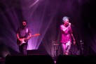Na scenie wokalistka i gitarzysta podczas występu. Scena oświetlona jest fioletowym światłem. W tle widać perkusję. 