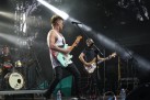 Zdjęcie przedstawia dwóch mężczyzn grających na gitarach. Z tyłu sceny jest również muzyk grający na perkusji. Widać sprzęt muzyczny np. w postaci mikrofonów. 