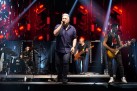 Na zdjęciu występ wokalisty ubranego na czarno i muzyków grających na gitarach i perkusji. Całość oświetla czerwone światło. 