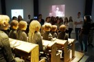 Na zdjęciu grupa młodych osób ogląda instalację artystyczną przedstawiającą manekiny dzieci siedzących w szkolnych ławkach.