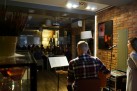 Zdjęcie przedstawia występ muzyczny w kameralnym wnętrzu. Widać na nim mężczyznę grającego na gitarze elektrycznej, siedzącego tyłem do wykonującego zdjęcie, kobietę stojącą obok niego, prawdopodobnie śpiewającą, z mikrofonem przed sobą, publiczność siedzącą przy stolikach w tle, słuchającą występu, a także sprzęt nagłośnieniowy i oświetlenie sceniczne.