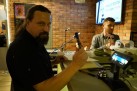 Na zdjęciu widać dwóch mężczyzn wewnątrz pomieszczenia restauracji. Na pierwszym planie mężczyzna z długimi włosami i brodą, ubrany w czarną koszulę, patrzy bezpośrednio w obiektyw. Trzyma w ręku długopis i kartkę papieru, opierając się o ladę. Na drugim planie za ladą znajduje się drugi mężczyzna, ubrany w szarą marynarkę. Za mężczyznami znajduje się ściana z czerwonej cegły. Widać na niej ekran, na którym widnieje napis. Na ladzie leży dzwonek i inne przedmioty.