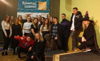 Na zdjęciu znajduje się grupa młodzieży pozująca razem na tle baneru programu „Równać Szanse”. Część uczestników ma na sobie codzienne ubrania, ale kilka osób przebranych jest w stroje nawiązujące do tematyki magicznej lub halloweenowej – m.in. czarownica w kapeluszu czy postacie w pelerynach. W tle widoczny jest baner projektu oraz drewniana i zielona ściana. - powiększ