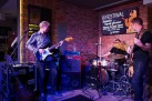Na zdjęciu dwaj mężczyźni grający na gitarach oraz mężczyzna grający na perkusji. Widać dużo sprzętu muzycznego, ceglane ściany oraz ekran z plakatem promującym odbywające się wydarzenie. Scena ma klimatyczny nastrój. 