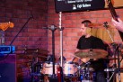 Na zdjęciu widać mężczyznę grającego na perkusji. Widać także część dwóch gitar. Za perkusistą znajduje się ceglasta ściana. 