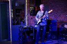 Na zdjęciu mężczyzna grający na gitarze elektrycznej. Widać sprzęt muzyczny. Światła są przyciemnione, co nadaje klimatu zdjęciu. 