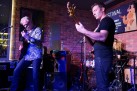 Dwóch mężczyzn grających na gitarach. Wokół nich sprzęt muzyczny. 