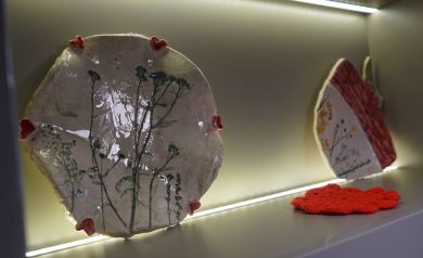Zdjęcie przedstawia ceramiczną płytkę, nieregularnego kształtu, ozdobioną wzorami roślinnymi (prawdopodobnie suszonymi roślinami wtopionymi w szkliwo) oraz małymi, czerwonymi serduszkami na brzegach. Drugi element dekoracyjny, również ceramiczny jest częściowo widoczny, z widocznymi czerwonymi i białymi powierzchniami oraz delikatnymi zdobieniami. Pod drugim elementem ceramicznym leży mały, czerwony przedmiot, wyglądający na wykonany szydełkiem lub na drutach, przypominający serwetkę lub inną drobną ozdobę. - powiększ
